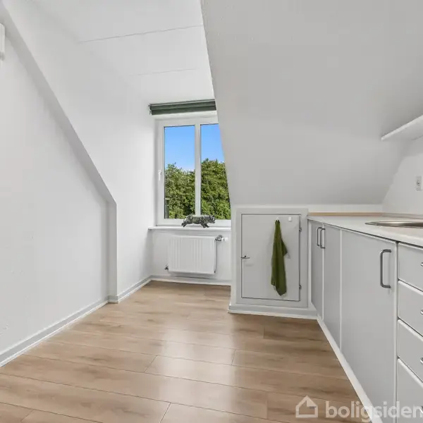 Et hvidt køkken med en række skabe og en håndvask. En grøn klud hænger på skabsdøren. Rummet har et skråt loft og et vindue med udsigt til træer.