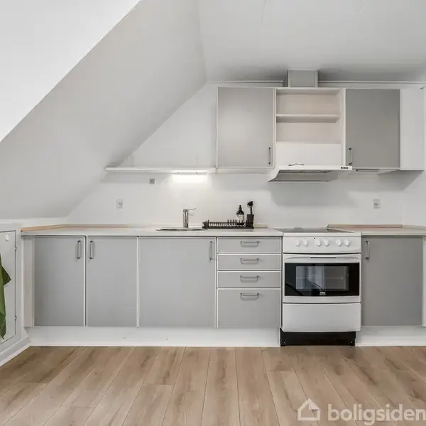 Et køkken med grå skabe og skuffer, hvor en ovn og vask er integreret. Rummet har skrå loft, lyse trælaminatgulve og en grøn klud hænger ved en skabsdør.