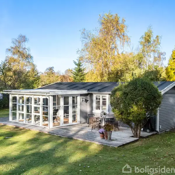 Et gråt sommerhus står på en række græsplæne omgivet af træer. Huset har en stor glasudestue og en træterrasse med havemøbler.