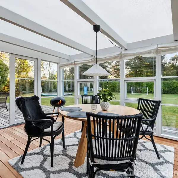En rund træbord med fire sorte stole står på et tæppe. En pendellampe hænger over bordet i en lys udestue med store vinduer, udsigt til have med græs og træer.