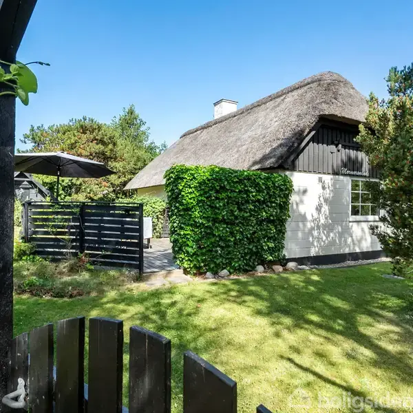 Stråtækt hus står omgivet af frodig grøn have med træer og hegn. Parasollen og terrassen er synlig til venstre. Klar blå himmel i baggrunden.
