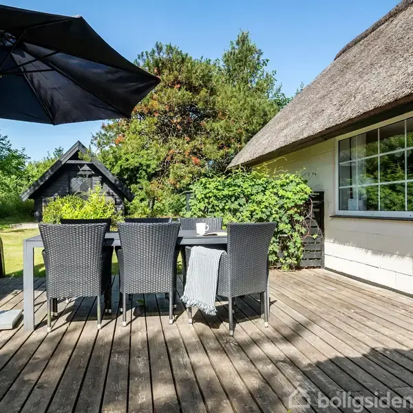 Terrassebord med fire flettede stole står under en stor sort parasol. Omgivet af frodige planter og et stråtækt hus i baggrunden, giver en hyggelig udendørs atmosfære.