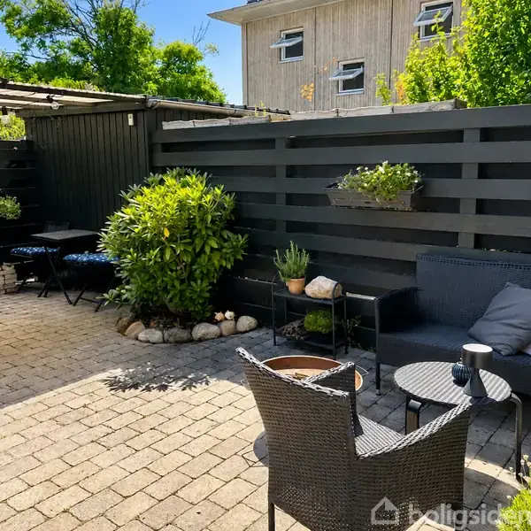 En udendørs terrasse med kurvemøbler og et lille bord står ved en sort trævæg. Planter og blomster dekorerer området. Baggrunden viser en større bygning og grønt træ.