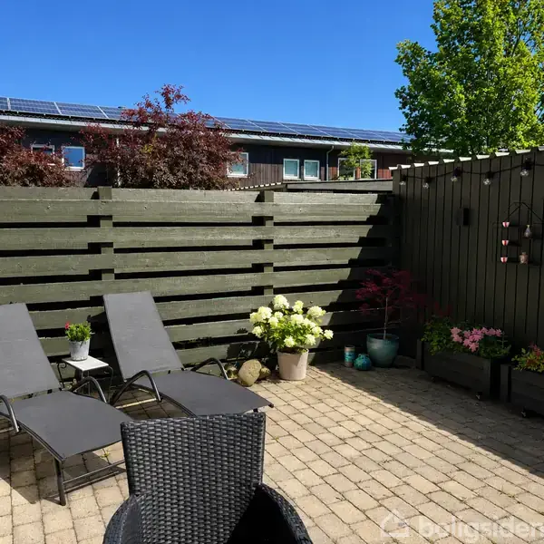 To liggestole står på en flisebelagt terrasse med drivhuse og blomster i krukker ved et plankeværk. Himlen er klar blå, og solen skinner over træer og buske.