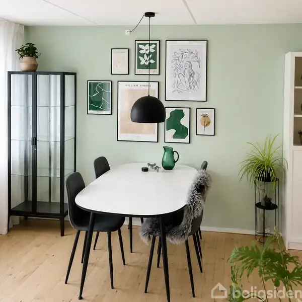 En hvid spisebord med sorte stole, dekoreret med en grøn kande og lys, står i et rum med lys grøn væg og kunstneriske plakater. En glasvitrine og plante til venstre.