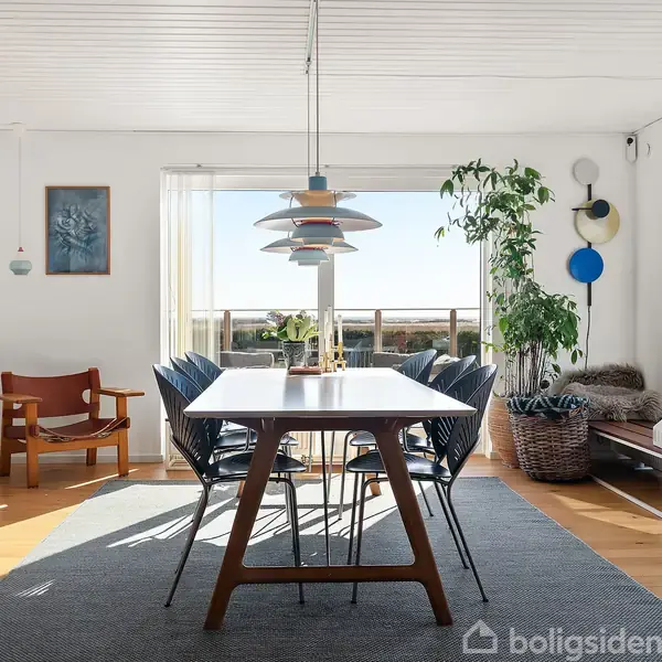 Spisebord med stole står i et lyst rum. En lampe hænger over bordet. Store vinduer lader sollys komme ind. Planter og dekorationer omgiver rummet.