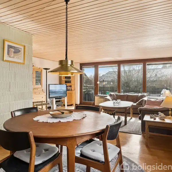 Spisebord med stole står i forgrunden, omgivet af en hyggelig stue med sofaer og et stort vindue, der giver udsigt til en have. En lampe hænger over bordet.