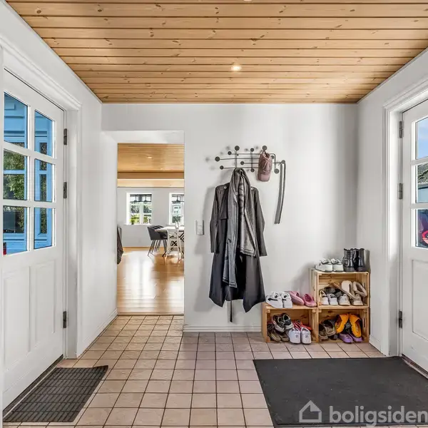 En entre med flisegulv har jakker hængende på en knagerække og sko på en hylde. To døre med glasvinduer fører til udendørsområder. I baggrunden ses et lyst rum.