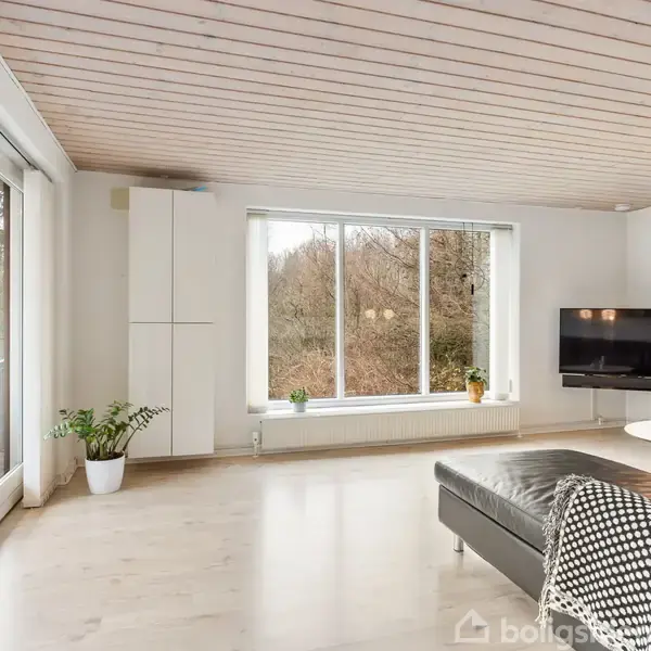 Stue med sorte lædermøbler, stor vinduesfront, der slipper naturligt lys ind, og fladskærms-tv på væggen. Trægulv og hvide vægge skaber en lys, moderne atmosfære.