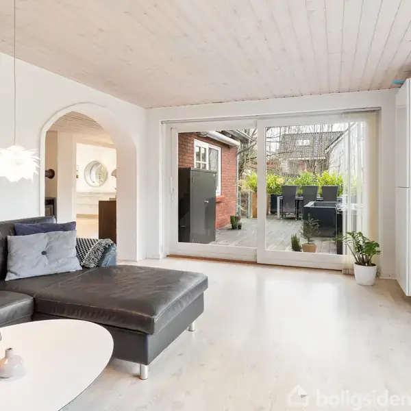 Sort lædersofa står i en lys stue med parketgulv, mens to pendellamper hænger fra loftet. Store vinduer åbner udsigt til en terrasse og planter udenfor.