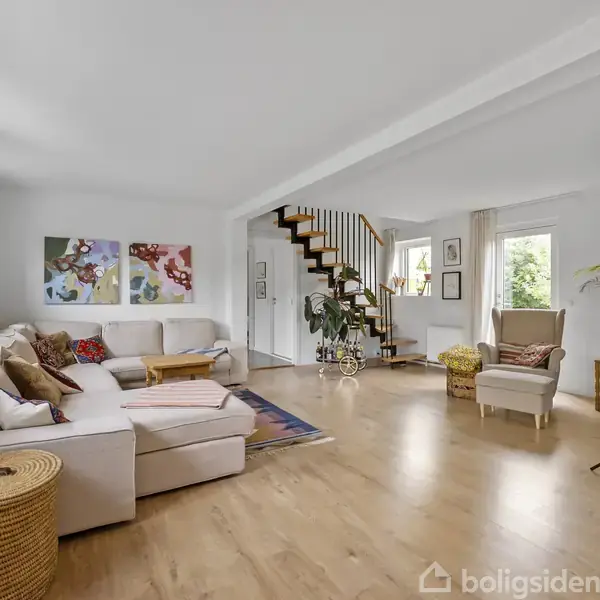 Stue med en stor hvid sofa, hvor der ligger puder og tæpper. Rummet har trægulv, vægkunst, planter og en trappe fører opad. Der er et stort vindue og lys indretning.