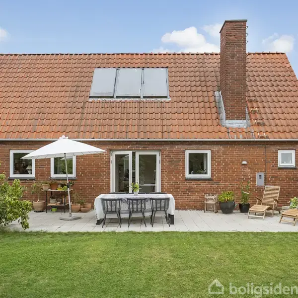 Rødt murstenshus med skråt tegltag har en terrasse med parasol, spisebord og stole. Omkring er en velplejet græsplæne og blomsterbede under en blå himmel med få skyer.