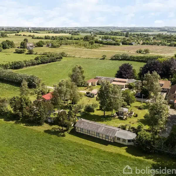 Et stort landligt område med en gård omgivet af træer og marker. Flere bygninger og et drivhus er synlige, mens marker strækker sig ud i det fjerne under en blå himmel.