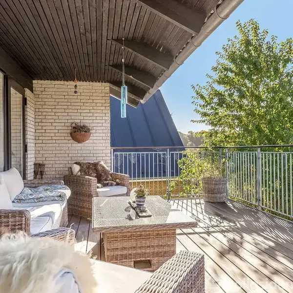 En møbleret terrasse med flettede sofaer og bord. Terrassebrædderne er træ, og der er en plantekrukke og hængende dekoration. Sollys skinner ind, omgivet af grønne træer.
