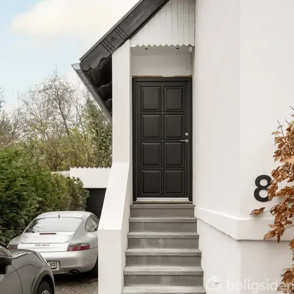 En sort hoveddør med paneldesign fører op fra en kort trappe. Til venstre holder en sølvfarvet bil ved grønne buske. Til højre ses et tal: "8".