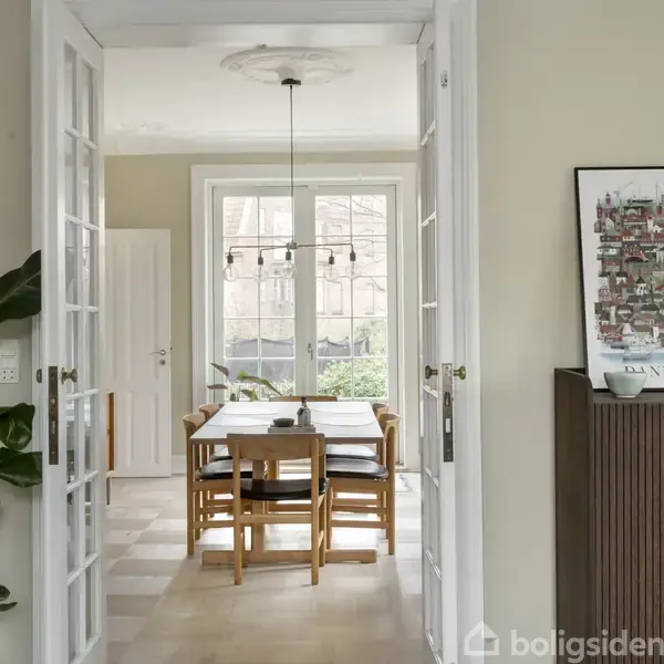 Spisestue med træbord og seks stole placeret under en pendellampe. Store vinduer bringer lys ind. En plante står til venstre, og en plakat er placeret over et skab til højre.