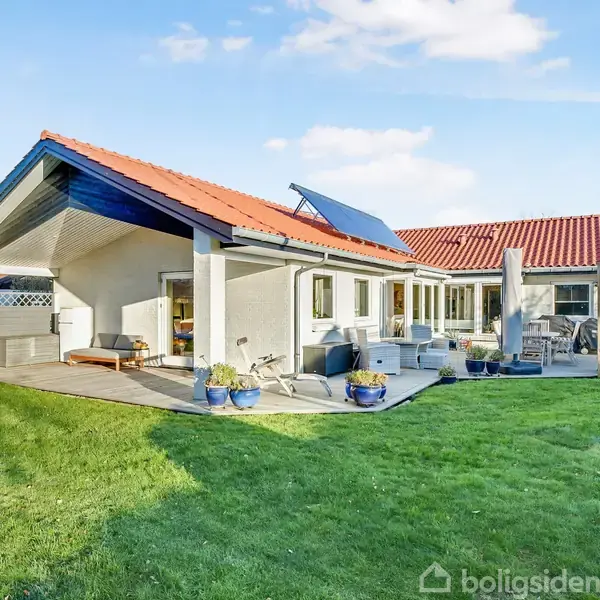 Hus med rødt tag og solpanel, omgivet af grøn græsplæne. Havemøbler står på en terrasse, mens krukker med planter dekorerer området. Skur ses i baggrunden.