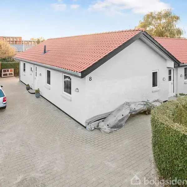 Hvidt hus med rødt tegltag står på brolagt gårdsplads. En bil er parkeret til venstre ved huset. Grønt hegn omkranser dele af gårdspladsen. Nummer 8 er på husvæggen.