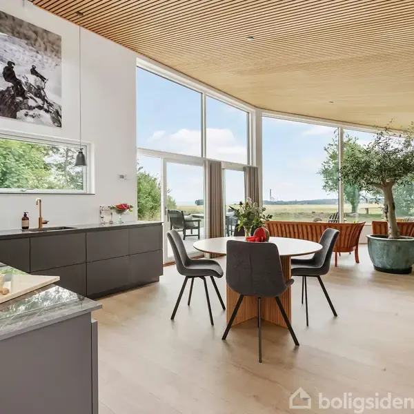 Køkken med spiseplads, bord omgivet af stole, brød på køkkenbordet, store vinduer med udsigt til terrasse og natur. Loft i træ, stor plante i hjørnet, indretning i moderne stil.