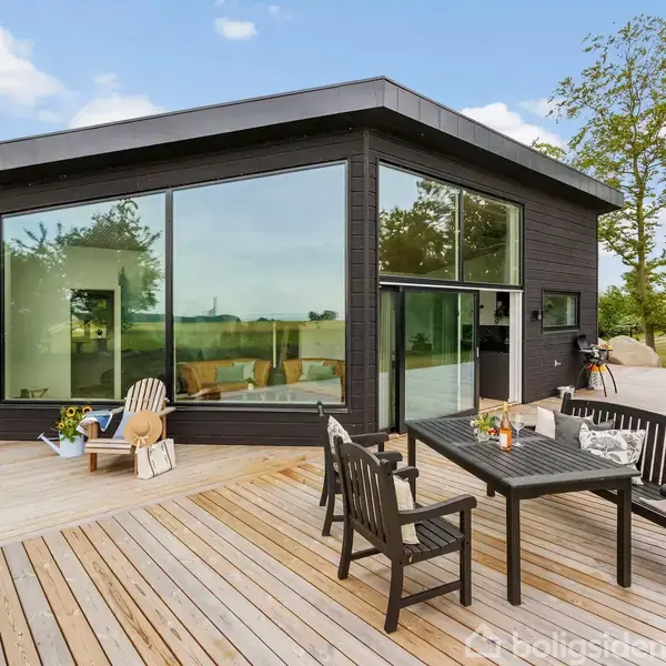 Et moderne sort hus med store glasvinduer står på en træterrasse. Et bord og stole er placeret udenfor, omgivet af grønne træer og en blå himmel i baggrunden.