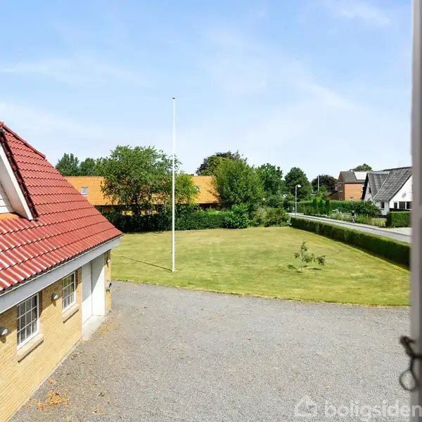 Bygning med rødt tegltag ses fra en åben vindue, udsigt til grusbelagt gårdsplads og græsplæne med flagstang i en rolig, grøn villakvarter.