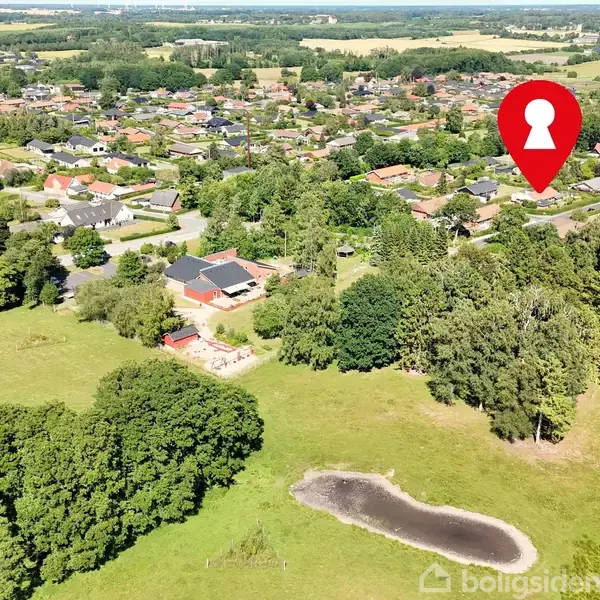 Landsby set fra luften med huse omgivet af træer. En rød markør med nøglehul indikerer en specifik placering. En mark med lille sø i forgrunden.