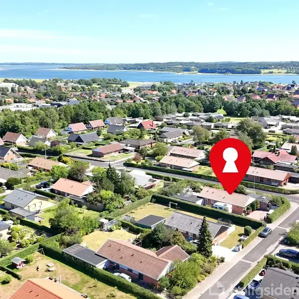 Et rødt ikon med en nøglesymbol markerer en ejendom i et boligområde. Omkringliggende huse med haver ses i en frodig, grøn landskab nær en stor sø i baggrunden.