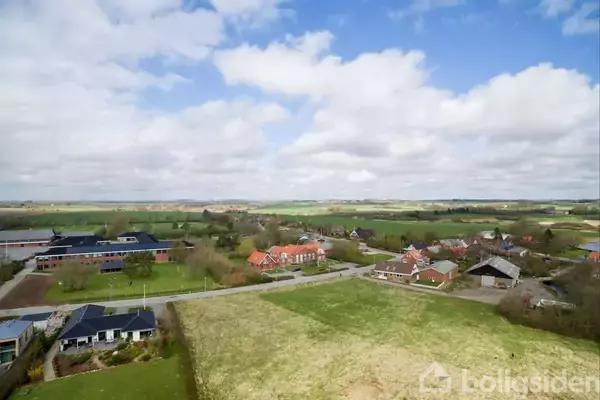 Landskab af marker og huse, set fra en høj vinkel. Flere bygninger ligger spredt i en lille landsby omgivet af åbne marker, under en delvist skyet himmel.