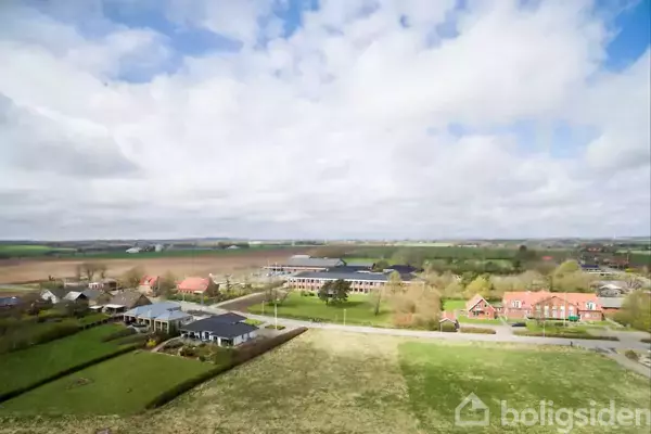 Landsbyhuse ligger spredt omkring en åben mark. Baggrunden domineres af store marker under en skyet himmel. Flere huse har røde tage, og området er omgivet af grønne områder.