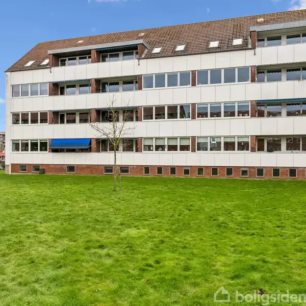 En moderne lejlighedsbygning med flere etager og store vinduer står foran en grøn græsplæne i et byområde. Klatrende planter ses nær murstensfundamentet.