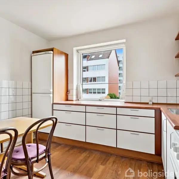 Et lyst køkken med brunt og hvidt interiør; et spisebord og stole står i midten, mens skabe og vask flankerer en vindue med udsigt til en bygning.