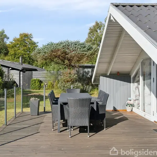 Terrassebord og stole står på en træterrasse ved et moderne sommerhus. Terrassen er omgivet af glasværn og græsplæne, med træer og buske i baggrunden under en blå himmel.