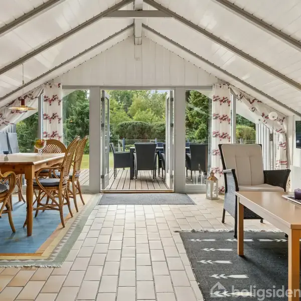 Et rummeligt, lyst rum med træmøbler og spisebord arrangeret til venstre, stueområde til højre. Store glasdøre åbner ud mod terrasse og grøn have.