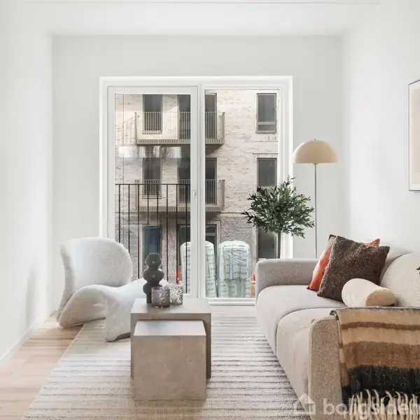 Stue med sofa og lænestol står ved siden af et lavt bord. En lampe og en plante er placeret ved siden af. Ud over glasdøren anes en bygning udenfor.