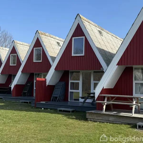 Røde A-formede hytter står på en græsplæne. Hytterne har store vinduer, og der ses udendørs møbler langs verandaerne. Himlen er klar og blå.