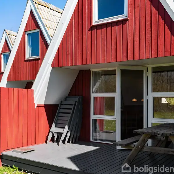 Røde, trekantede sommerhuse med hvide vinduer står i række på en græsplæne. Foran huset ses et brunt træbord med bænke på en terrasse. Et skilt viser nummeret "B305".