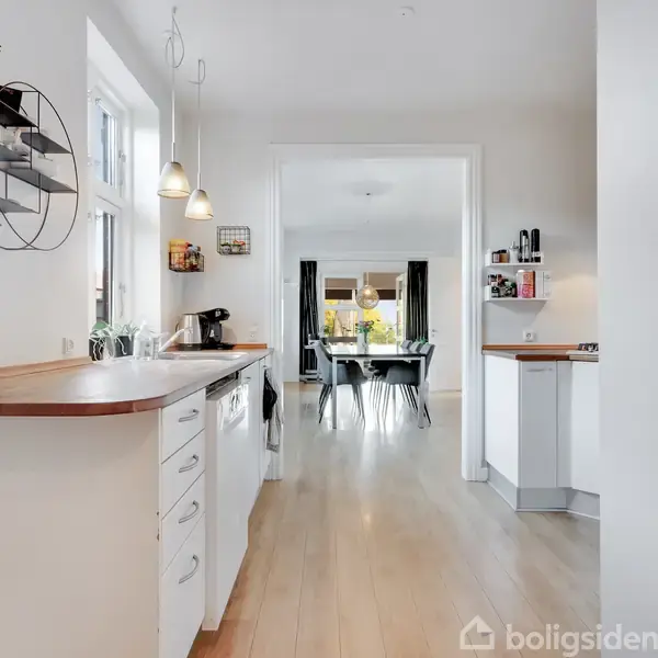 Et køkken med hvide skabe og træbord, en kaffemaskine og hængende lamper. I baggrunden ses en spisestue med et bord og stole ved et vindue.
