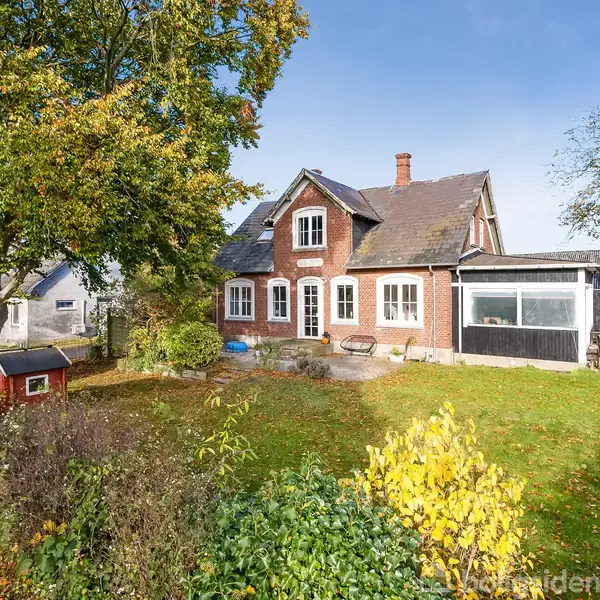 Et rødt murstenshus står i en have med grønne træer og en lille rød legehus. Paraplyformede træer omkranser ejendommen under en klar blå himmel.