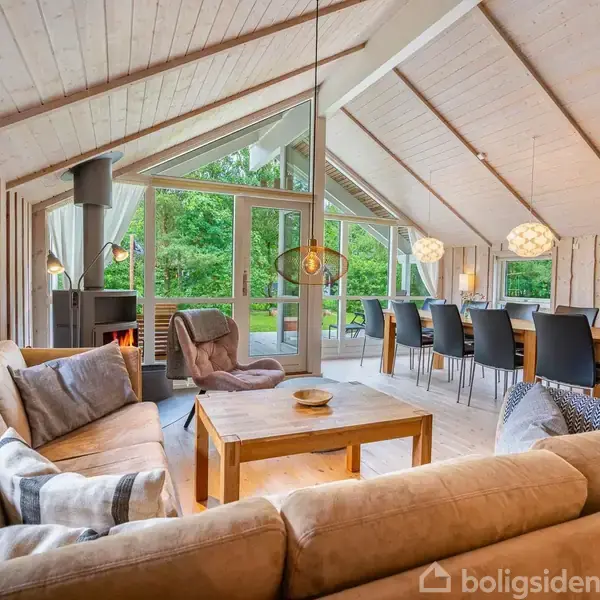Stue med beige hjørnesofa og træbord, pejs i brug. Store vinduer med udsigt til frodig have. Langt spisebord med sorte stole, moderne loftlamper.