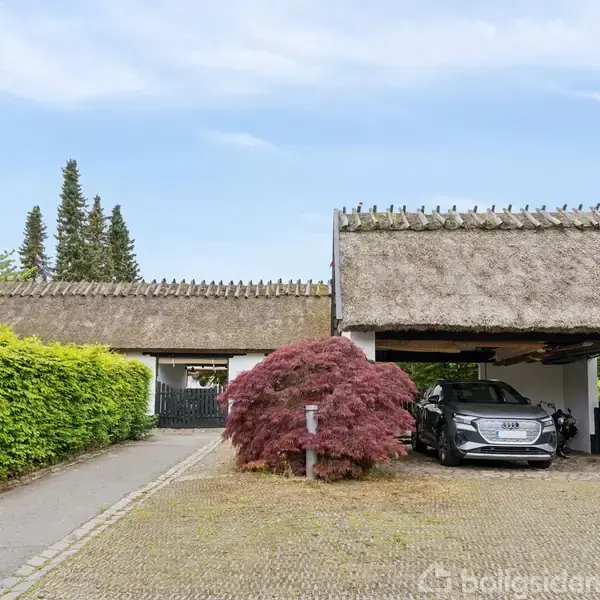 En bil holder parkeret under et stråtækt carport-tag. Til venstre ses en sti flankeret af grønne buske og et lille træ. Huset har hvid mursten og er beliggende i et grønt område.