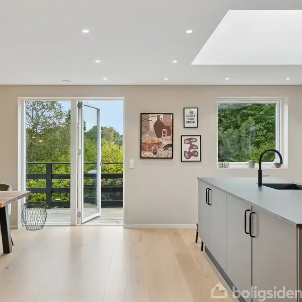 Et moderne køkken med en grå køkkenø, sorte greb, og en vandhane. Bordet har sorte stole omkring sig. Store vinduer og en åben dør giver udsigt til grønne træer.