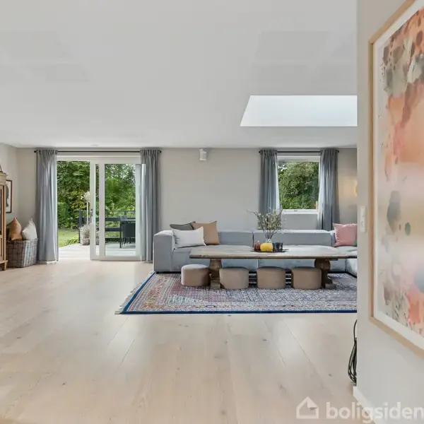 Et stueområde indeholder en stor, grå sofa og lavt sofabord på et orientalsk tæppe. Store vinduer med grå gardiner slipper udsigt til grøn have ind. Farverig kunst hænger på væggen.