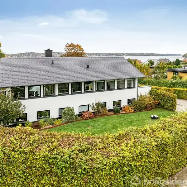 Et hvidt hus med sort tag står i en have omgivet af en hæk. I baggrunden ses træer og vand. Huset har store vinduer og en velplejet græsplæne foran.