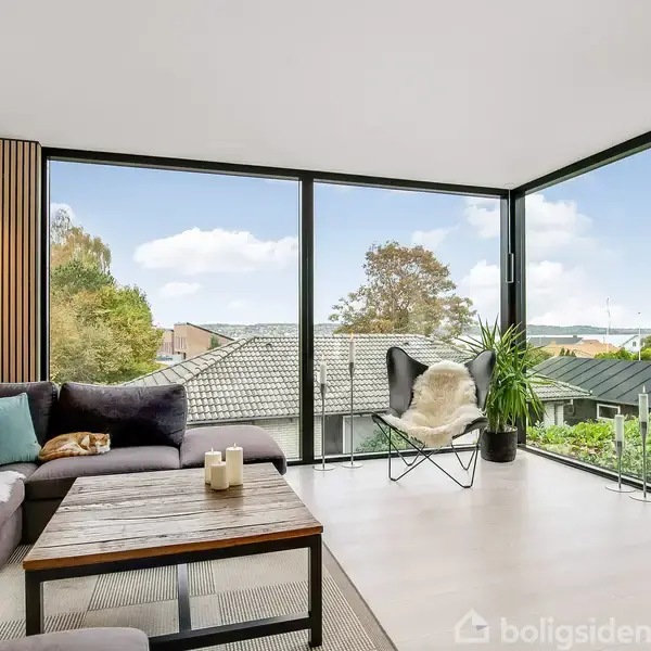 Stue med en L-formet sofa, bord og et teleskop ved et stort hjørnevindue. Udsigt til hustage og træer udenfor. Et lille bord med grønne planter skaber en moderne atmosfære.