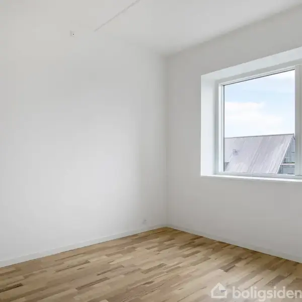 Et tomt rum med trægulv har en stor vinduesramme, der slipper lys ind. De hvide vægge skaber et lyst og rummeligt indtryk.