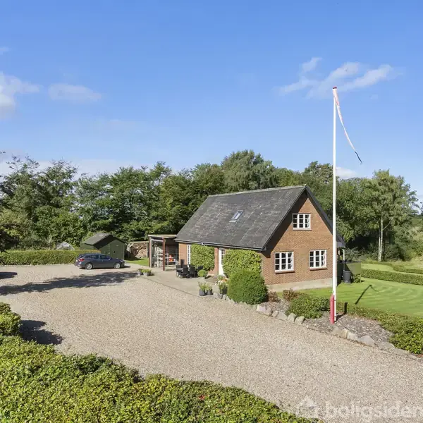 Et rødt murstenshus med hældningstag står i en grøn have. Til venstre en parkeret bil på gruset vej. Flagrende flag i forhaven omgivet af træer og buske.