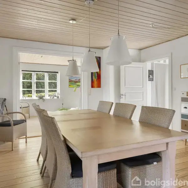 Et stort spisebord med syv fletstole står i et lyst spisestueområde. Ovenover hænger tre hvide lamper. I baggrunden ses en åben dør og et vindue.