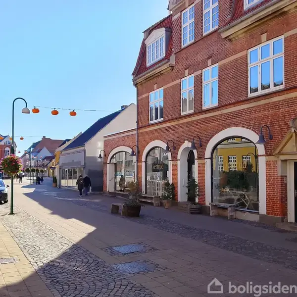 Rød murstensbygning med buede vinduer står langs en brostensbelagt gågade. To mennesker går forbi butikker. Hængende lamper og blomster skaber en hyggelig byatmosfære under blå himmel.