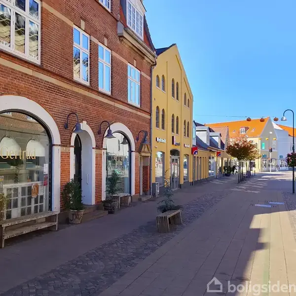 Butikkerne med murstensfacader og buede vinduer står langs en stille gågade med brosten og blomsterkurve, hvor solen skinner klart på bygningerne. En skilt viser teksten "Beth Baun".