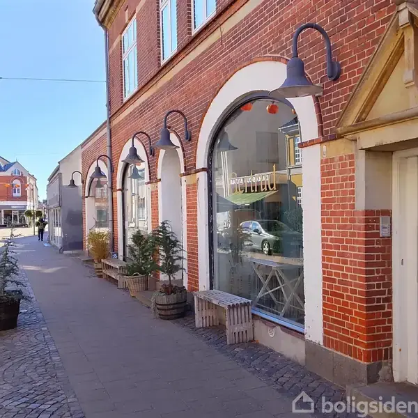 Røde murstensbygninger med buede vinduer og butikker. Udendørs planter placeret langs facaden. Gadelamper hænger over en brostensbelagt fortov. En dør markeret "4" er synlig til højre.